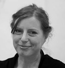 Eine Frau mit heller Haut und zurückgebundenem hellem Haar und einem dunklen Oberteil lächelt vor einem schlichten Hintergrund in die Kamera.