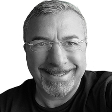 Ein Mann mittleren Alters mit kurzen grauen Haaren, Brille und Bart lächelt vor einem schlichten weißen Hintergrund in die Kamera.