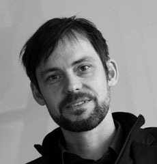 Ein Mann mit kurzen dunklen Haaren, Bart und Schnurrbart blickt in die Kamera. Er trägt ein dunkles Hemd mit Kragen und eine Jacke vor einem schlichten Hintergrund.