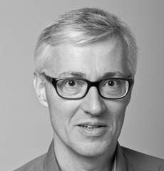 Ein Mann mit kurzen grauen Haaren und einer schwarz umrandeten Brille sowie einem Hemd mit Kragen blickt vor einem schlichten Hintergrund in die Kamera.