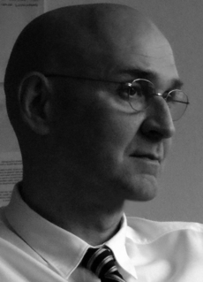 Ein Mann mit Brille und Glatze trägt ein Hemd mit Kragen und gestreifter Krawatte und blickt zur Seite. Das Bild ist schwarz-weiß.