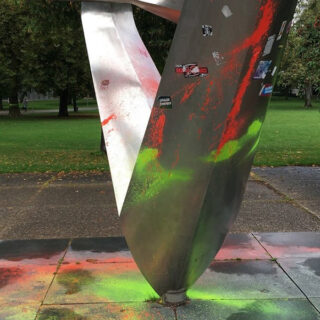 Eine Metallskulptur auf einer gepflasterten Fläche in einem Park ist mit grüner und orangefarbener Farbe bespritzt und mit Aufklebern versehen. Im Hintergrund sind Bäume und Gras zu sehen.