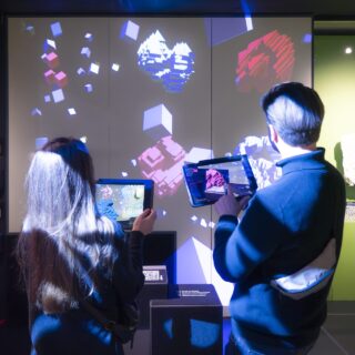 Zwei Personen mit Tablets interagieren mit einer auf eine Wand projizierten digitalen Kunstanzeige mit bunten geometrischen Formen und Würfeln.