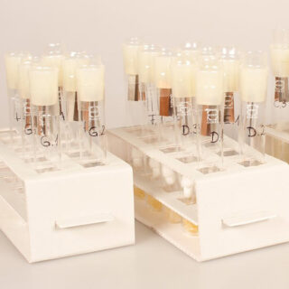 Eine Gruppe von Reagenzgläsern mit weißen Stopfen ist in zwei weißen Gestellen angeordnet. Jedes Reagenzglas ist mit einem schwarzen Stift beschriftet und enthält am Boden eine kleine Menge Flüssigkeit.