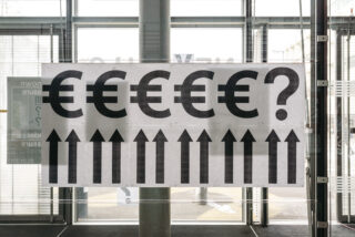 Auf einem Schild sind Reihen von Eurosymbolen und ein Fragezeichen über einer Reihe nach oben zeigender Pfeile zu sehen, was auf Fragen zu steigenden Werten oder Preisen des Euro hindeutet.