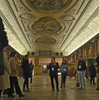 Eine Gruppe von Menschen hört einem Führer in einer großen Halle mit verzierten Decken und historischen Kunstwerken zu; an den Seiten des Raumes sind Kutschen zu sehen.