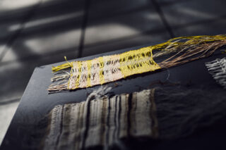 Nahaufnahme von gewebten Textilmustern mit losen Fäden, eines davon in Gelb- und Beigetönen, platziert auf einer dunklen Oberfläche mit Fliesenboden im Hintergrund.