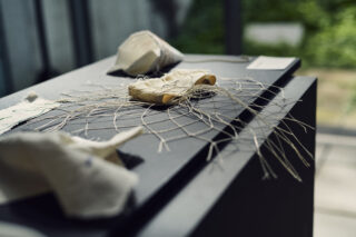 Eine Nahaufnahme eines textilen Kunstwerks mit gewebten Fäden und Stoff auf einer schwarzen Oberfläche, mit verschwommenen Stoffstücken im Hintergrund.