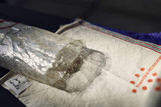 Eine Nahaufnahme einer Rolle aus silbernem, netzartigem Stoff auf einem cremefarbenen Textil mit roter und blauer Stickerei und einem teilweise sichtbaren handgeschriebenen Etikett.