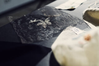 Ein durchscheinendes Stück schwarzen Stoffs mit einem silbern gestickten Blumenmuster liegt zwischen anderen Textilmustern auf einem Tisch.