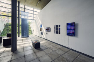 Eine moderne Kunstgalerie mit verschiedenen digitalen Bildschirmen, Fotografien und Kunstwerken an einer weißen Wand, die durch große Fenster mit natürlichem Licht beleuchtet wird.