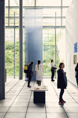 Menschen stehen und gehen in einer modernen Kunstgalerie mit großen Fenstern umher und betrachten Kunstwerke an den Wänden und einem Tisch in der Mitte.