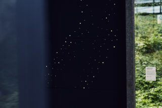Eine schwarze Platte mit kleinen quadratischen Löchern, die an Sterne erinnern, ist neben einem Fenster angebracht, durch das man draußen Grünpflanzen sieht und auf dessen Glas ein Schild angebracht ist.