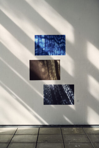 Drei abstrakte Fotografien in Blau- und Brauntönen sind vertikal an einer weißen Wand angebracht, wobei das Sonnenlicht diagonale Schatten über die Szene wirft.