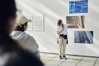 Eine Frau mit einem Mikrofon zeigt auf digitale Kunstausstellungen an einer Galeriewand, während sie zu einem Publikum spricht.