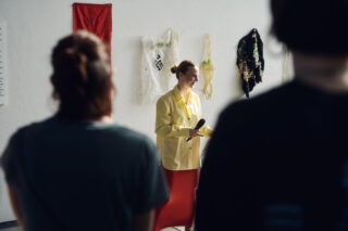 Eine Person in einem gelben Hemd steht mit einem Mikrofon vor textilen Kunstwerken an einer Wand, im Vordergrund sind zwei Personen zu sehen.