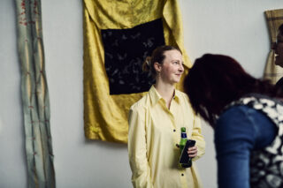 Eine Person in einem gelben Hemd hält eine Flasche und steht in der Nähe von Wandbehängen, während zwei andere im Vordergrund interagieren.