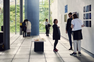 In einem modernen Galerieraum mit großen Fenstern, hohen Säulen und Kunstwerken an den Wänden betrachten Menschen Kunstwerke und unterhalten sich.