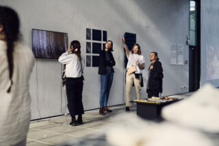 Vier Personen beobachten und diskutieren digitale Kunst, die auf Bildschirmen und Tafeln in einem modernen Galerieraum angezeigt wird.