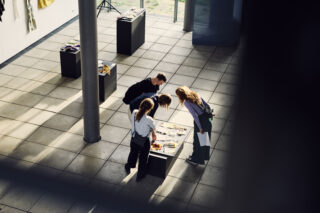 Vier Personen stehen in einem modernen Galerieraum mit gefliesten Böden und verstreuten Podesten um einen Ausstellungstisch herum und betrachten Materialien oder Kunstwerke.