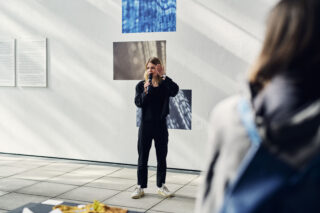 Eine Person in dunkler Kleidung spricht vor ausgestellten Kunstwerken und Texten auf einer weißen Wand in ein Mikrofon und wendet sich an ein unsichtbares Publikum.