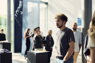 Ein Mann in einem grauen T-Shirt steht mit den Händen in den Taschen in einer hell erleuchteten Galerie, umgeben von anderen Menschen, die sich Ausstellungsstücke ansehen.