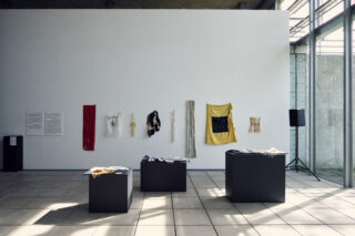 In einem minimalistischen Galerieraum hängen verschiedene Stoffe an einer weißen Wand, während im Vordergrund drei schwarze Sockel mit weiteren Textilstücken stehen.