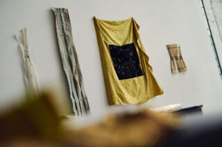 Stoffstücke in verschiedenen Farben und Mustern sind an einer schlichten weißen Wand drapiert und aufgehängt, wobei einige Falten und unregelmäßige Anordnungen sichtbar sind.