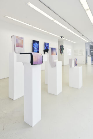 Eine Galerie für zeitgenössische Kunst mit weißen Ausstellungssockeln, auf denen aufgerollte Stoffkunstwerke stehen, digitalen Bildschirmen an der Wand und an der Wand montierten Informationstafeln.