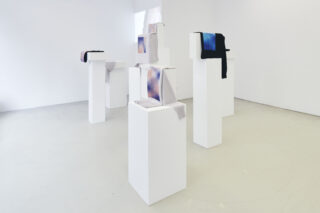 Auf weißen Podesten werden Stoffstücke mit Mustern in Blau-, Lila- und Schwarztönen in einem minimalistischen Galerieraum mit weißen Wänden und Böden ausgestellt.