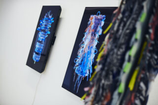 An einer weißen Wand hängen zwei digitale Kunstwerke mit abstrakten, farbenfrohen, pinselartigen Formen; eines wird teilweise von einem strukturierten Objekt im Vordergrund verdeckt.