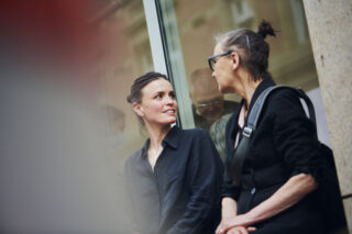 Zwei Frauen sitzen draußen auf einer Bank und unterhalten sich in der Nähe eines Gebäudes mit Glasfenstern.