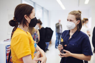 Zwei Frauen mit Gesichtsmasken unterhalten sich im Innenbereich; eine hält ein Getränk in der Hand. Andere Personen sind im Hintergrund verschwommen zu sehen.