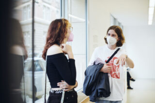 Zwei Menschen mit Mundschutz stehen drinnen an einem großen Fenster und unterhalten sich; einer hält eine Jacke in der Hand und gestikuliert mit der Hand.