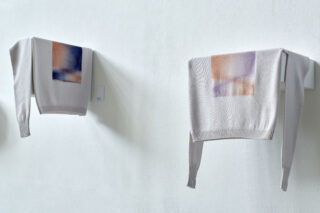 Zwei helle Pullover mit abstrakten Farbblöcken, die über weiße Wandregale vor einer blassen Wand drapiert sind.