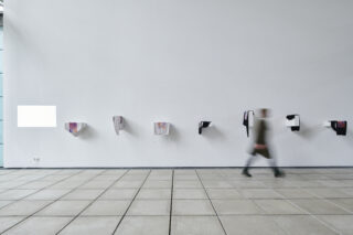 Eine Person geht an einer weißen Wand vorbei, an der mehrere abstrakte, kastenförmige Kunstwerke in einer Reihe über einem gefliesten Boden angebracht sind.