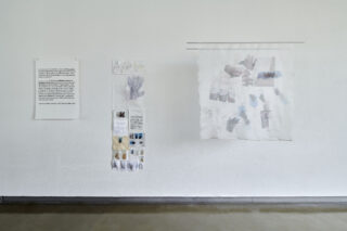 Kunstinstallation an einer weißen Wand mit einem bedruckten Textblatt, einem Textilstück mit verschiedenen kleinen Stoffmustern und einem hängenden durchscheinenden Stoff mit Handabdruck-Designs.