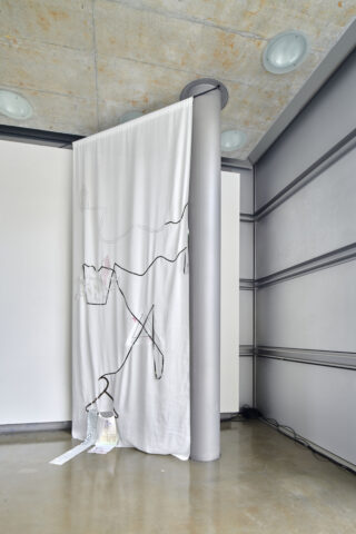Ein weißer Stoffvorhang mit abstrakter Strichzeichnung hängt an einer Stange, die an einer runden grauen Säule in einem modernen Raum mit Betondecke und poliertem Boden befestigt ist.