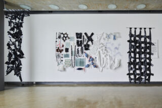 Eine weiße Galeriewand zeigt eine geordnete Anordnung verschiedener schwarzer und weißer skulpturaler Objekte und Materialien, flankiert von zwei vertikal hängenden schwarzen Strukturen.