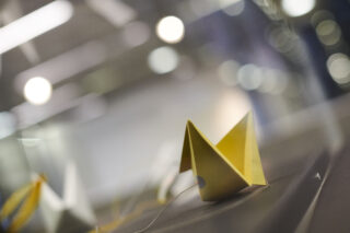 Ein gelbes Origami-Boot aus Papier liegt auf einer reflektierenden Oberfläche mit verschwommenem Licht und einem weichgezeichneten Hintergrund.