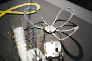 Ein Robotikprojekt mit einer blütenblattartigen Kunststoffstruktur, elektronischen Komponenten, Kabeln und einem gelben Maßband auf einer dunklen Oberfläche.