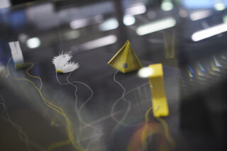 Nahaufnahme abstrakter Papierskulpturen mit geometrischen Formen und Ausschnitten, verbunden durch dünne Drähte, dargestellt auf einer reflektierenden Oberfläche mit unscharfem Hintergrund.