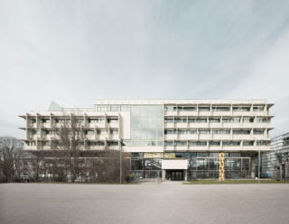 Ein modernes mehrstöckiges Gebäude mit Balkonen, großen Fenstern und einem zentralen Eingang; auf dem Rasen in der Nähe des Eingangs steht eine goldene Skulptur.