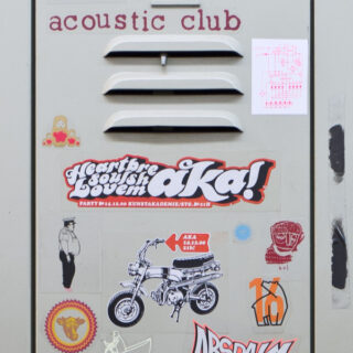 Eine beige Schließfachtür mit verschiedenen bunten Aufklebern, darunter Text, ein Motorrad und eine Comicfigur, unter einem Schild mit der Aufschrift „Acoustic Club“.