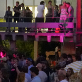 Bei einer Veranstaltung im Freien versammelt sich eine Menschenmenge; einige stehen auf einer erhöhten Plattform, während andere sich unten im Abendlicht unterhalten.