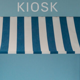 Blau-weiß gestreifte Markise, darüber steht in weißer Schrift auf blauem Grund „KIOSK“.