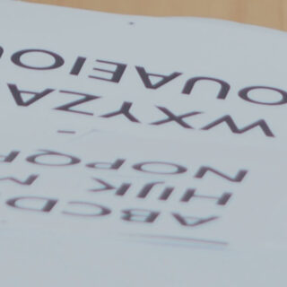 Auf einer Holzoberfläche sind zwei gedruckte Alphabettafeln gestapelt, die jeweils Großbuchstaben in Reihen darstellen.