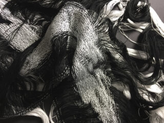 Schwarze und weiße Fäden sind verwoben und locker miteinander verwickelt, wodurch ein strukturiertes, abstraktes Muster auf dunklem Hintergrund entsteht.