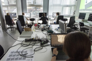 In einem modernen Büro arbeiten Menschen an Computern. Im Vordergrund entwirft eine Person auf einem Laptop ein 3D-Tiermodell. Auf dem Tisch stehen verschiedene VR- und Technikgeräte.