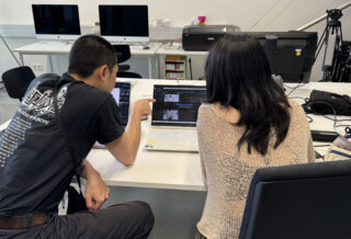 Zwei Personen sitzen an einem Schreibtisch, schauen auf Laptop-Bildschirme und diskutieren Inhalte in einem modernen Büro mit Computern und Elektronik im Hintergrund.